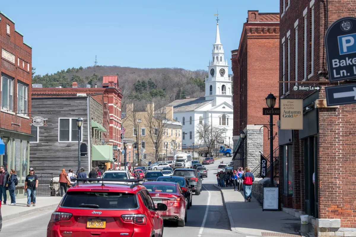 Bustling town street with cars and pedestrians leading towards a white church with a steeple.