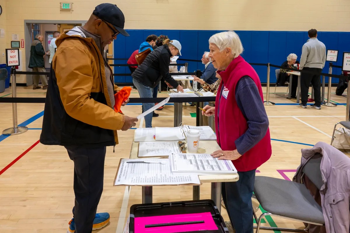 People are at a voting station inside a gymnasium, interacting with staff at tables.
