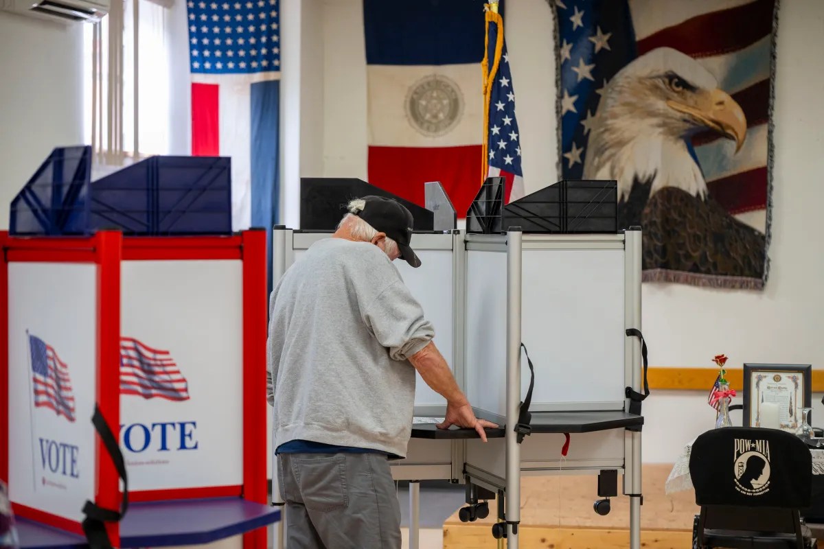 A person is standing at a voting booth inside a room decorated with American flags and an eagle tapestry.