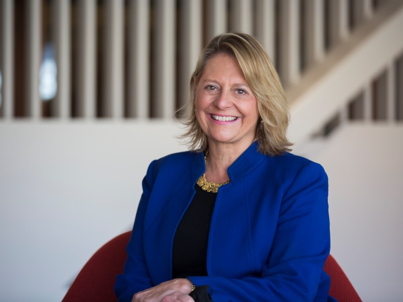 A woman with blonde hair wearing a blue blazer and black top sits on a red chair, smiling at the camera. There is a staircase with white vertical railings in the background.