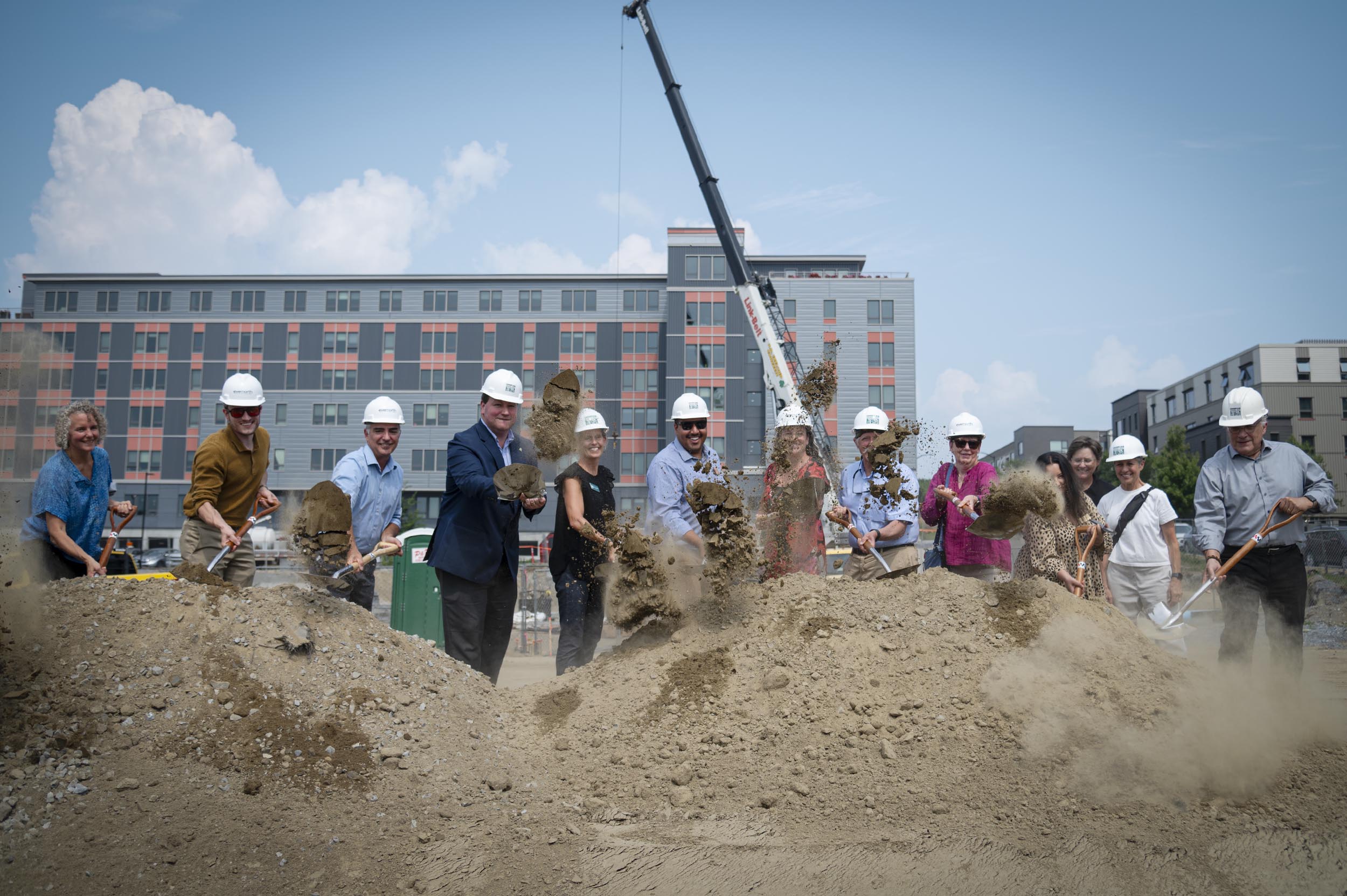A group of people in hard hats use shovels to break ground at a construction site in front of a modern building, with a crane in the background.