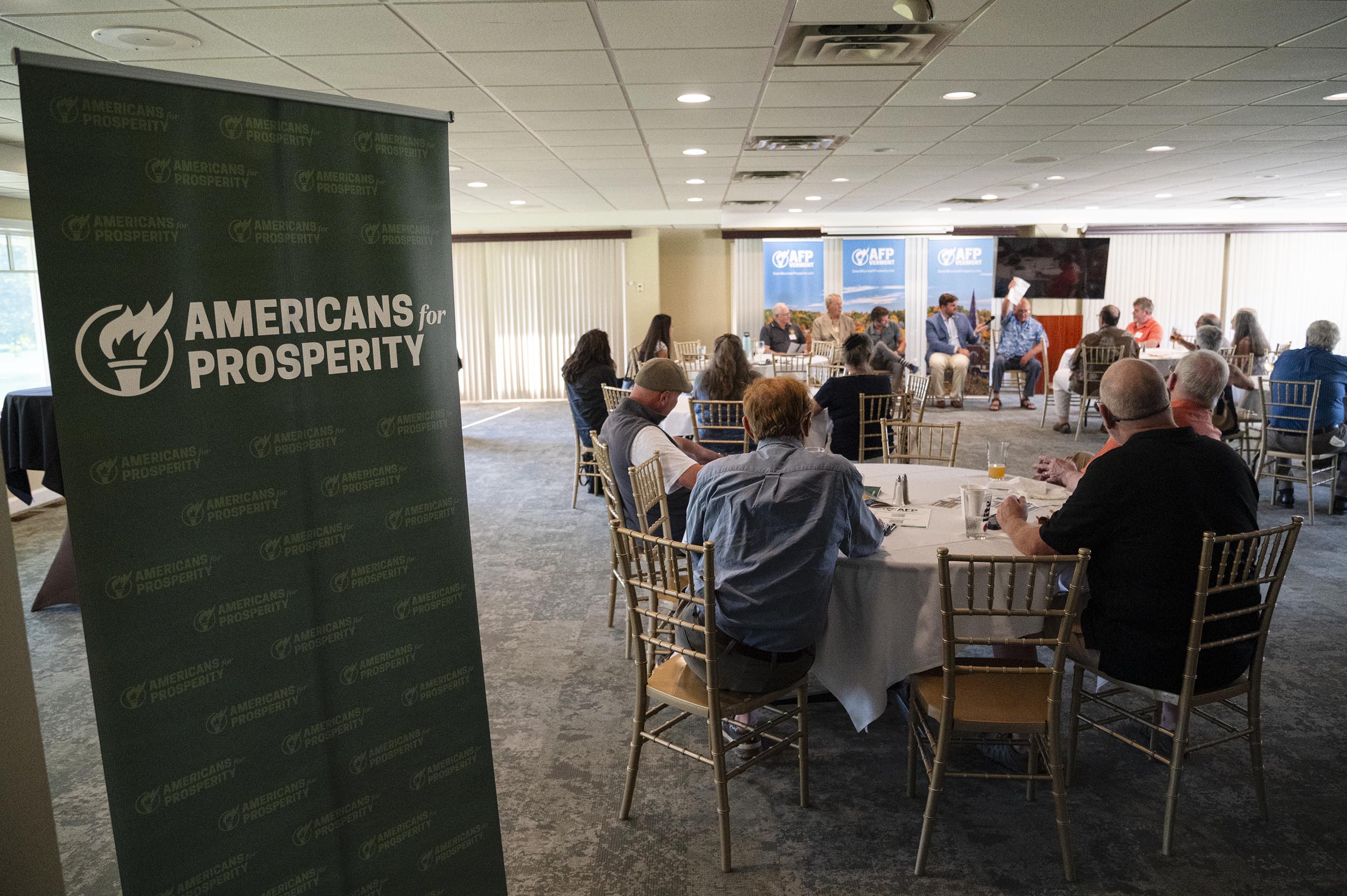 People sit at round tables listening to a panel discussion in a conference room; an "Americans for Prosperity" banner stands in the foreground.