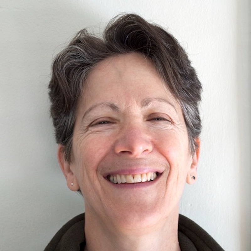 A person with short hair smiles at the camera, standing against a plain light-colored wall.