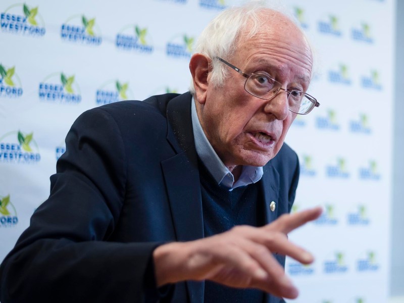 An older man wearing glasses and a dark suit gestures with his hand while speaking in front of an Essex Westford branded backdrop.