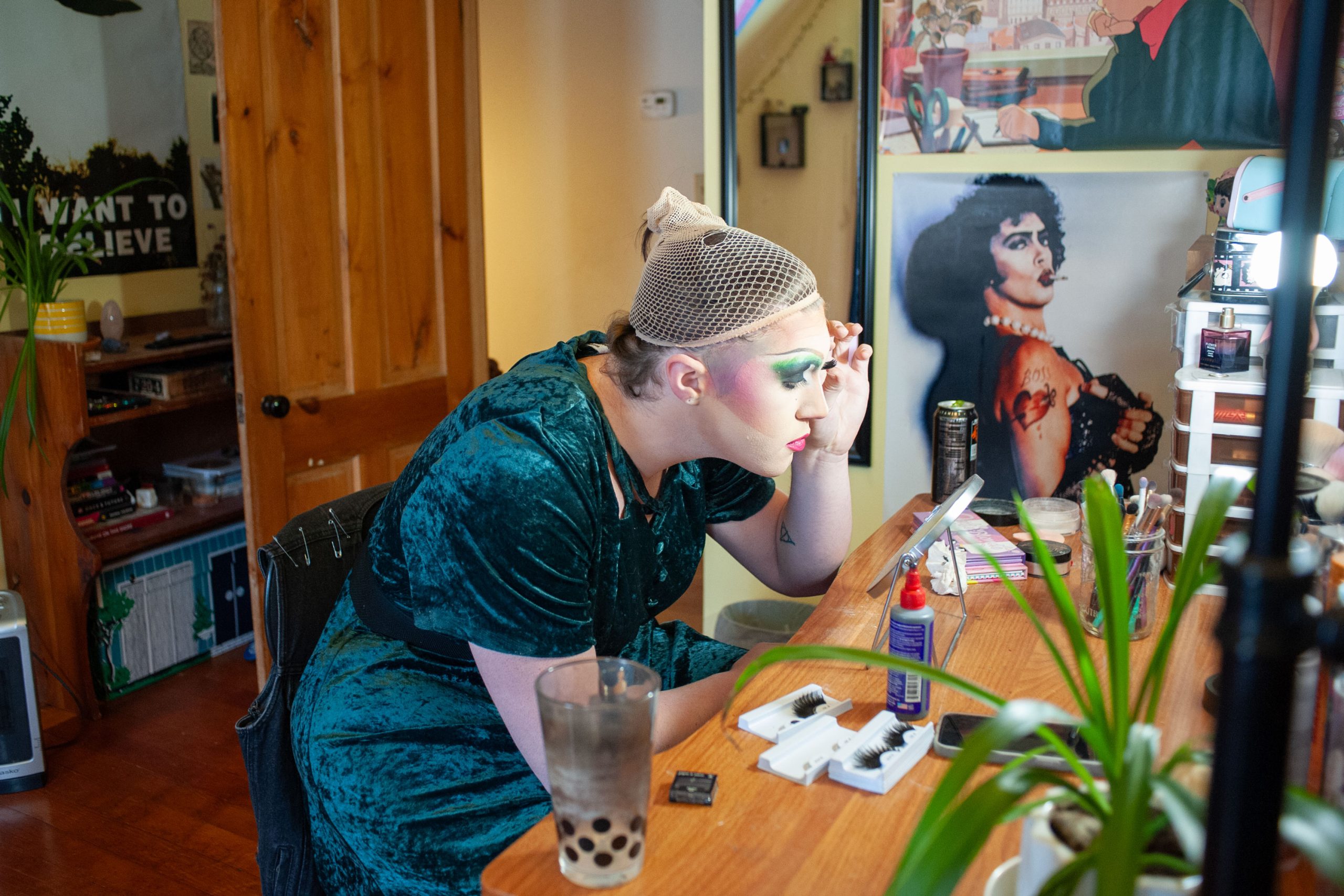 A person applies makeup at a dressing table, surrounded by beauty products, with a poster on the wall behind them.