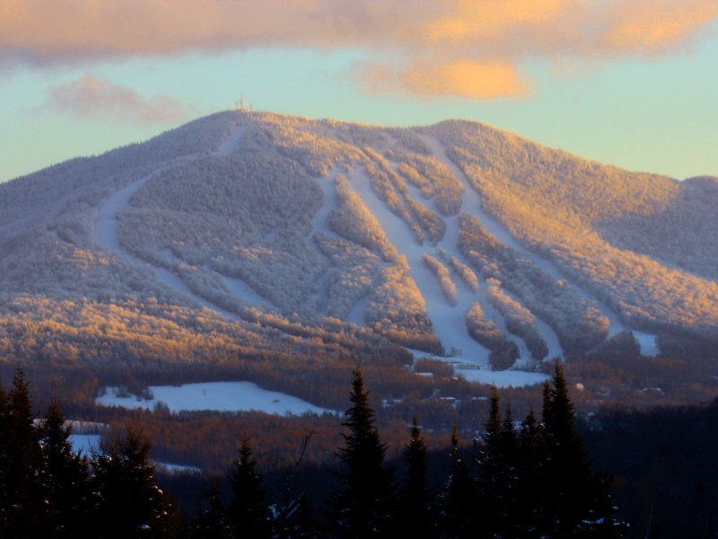 Snow-covered mountain with ski trails at sunset, surrounded by evergreen trees. Cloudy sky with orange hues.