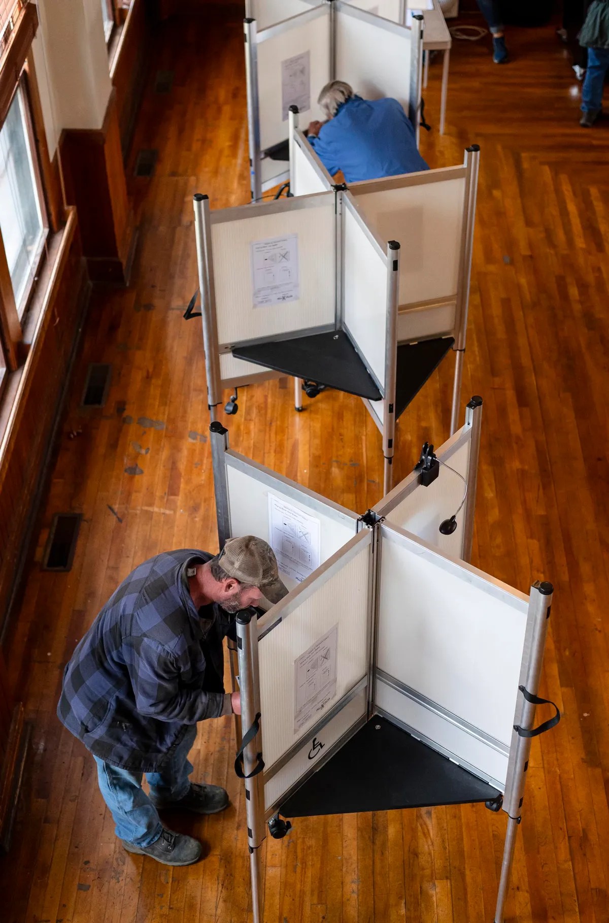 People voting in individual booths in a room with wooden floors, viewed from above.