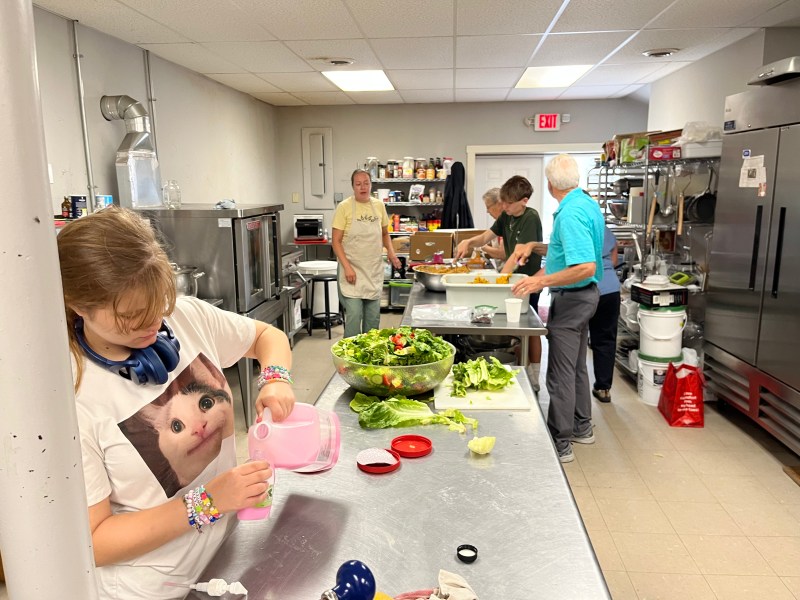 Five people work in a commercial kitchen preparing food; one pours liquid at the counter, while others chop vegetables. Large bowls of salad sit on the stainless steel workspace.