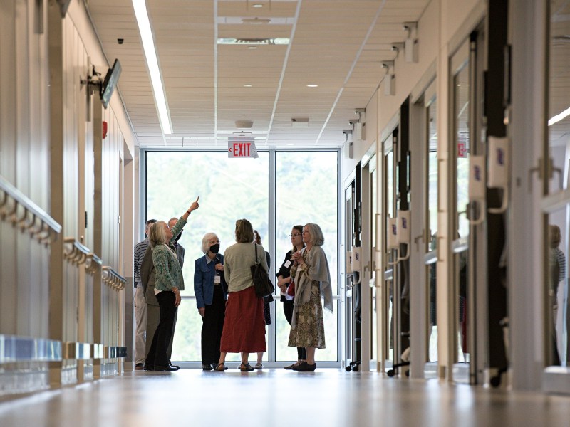 A group of six adults stands in a hallway, with one person pointing upward while others look on. The hallway has large windows at the end and doors on both sides.
