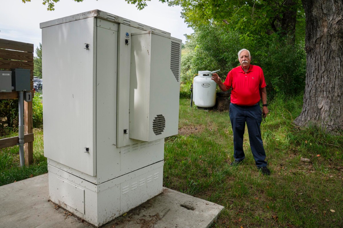 A man in a red shirt stands outdoors next to a white utility box, holding a propane tank in one hand.