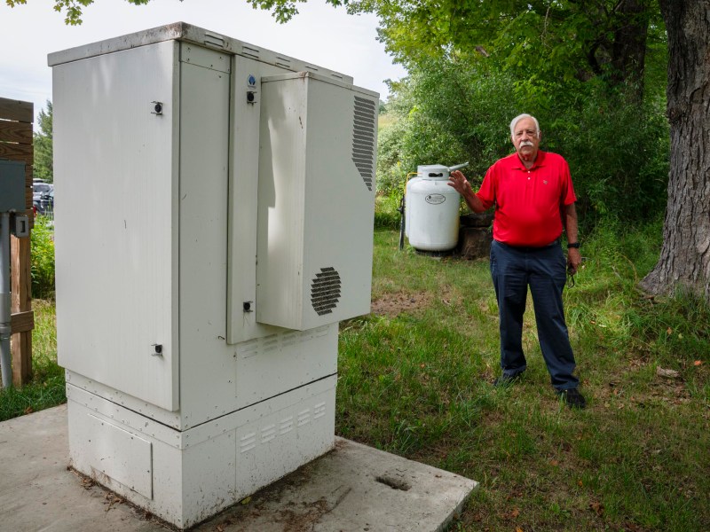 A man in a red shirt stands outdoors next to a white utility box, holding a propane tank in one hand.