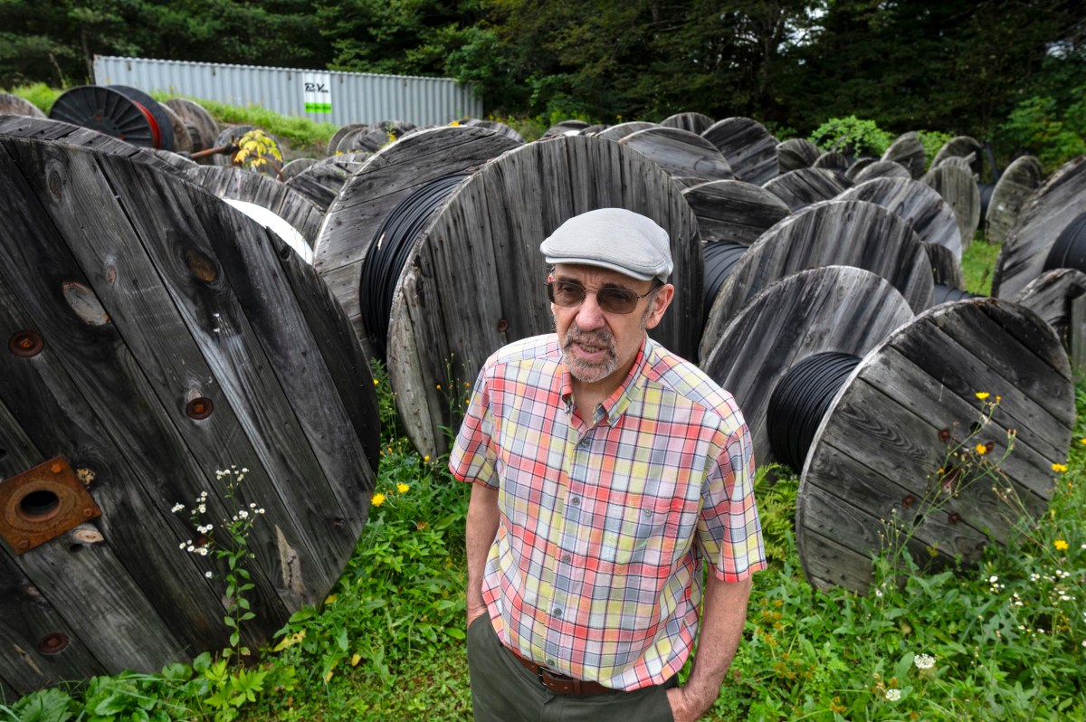 A man in a plaid shirt and cap stands among large wooden cable spools in a grassy outdoor area.