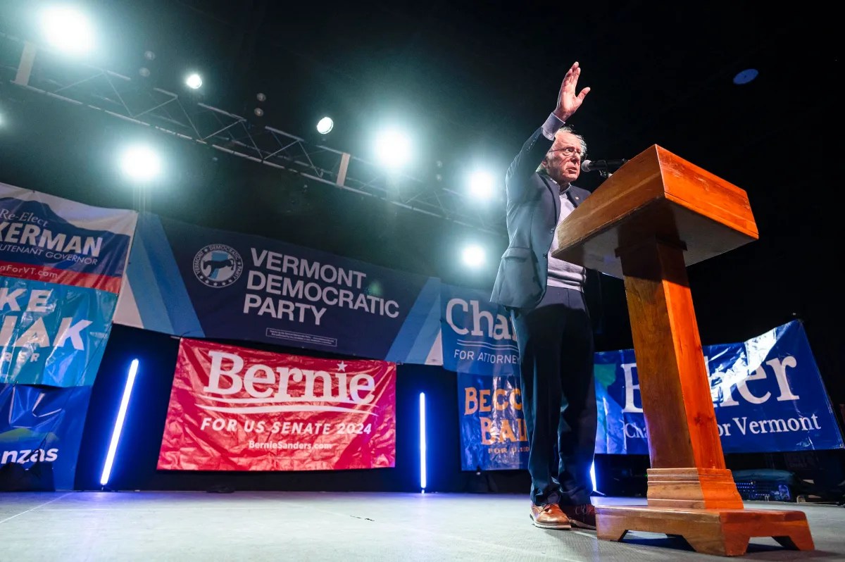 A person speaks at a podium during a Vermont Democratic Party event with banners and bright stage lights.