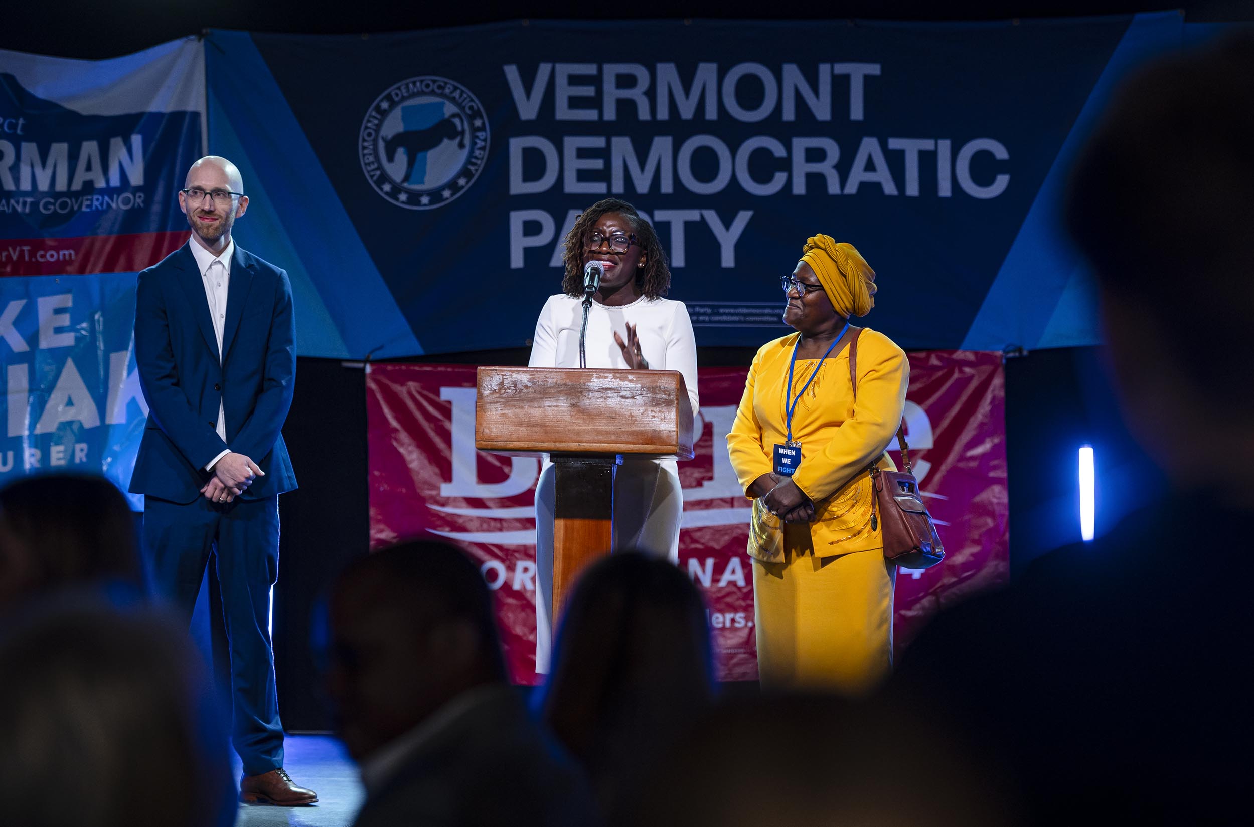 Three people on stage at a Vermont Democratic Party event, with one person speaking at a podium.