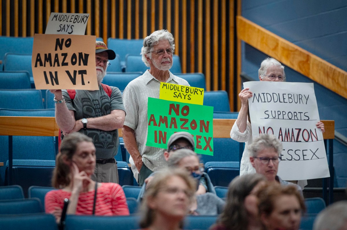 People at a meeting hold signs reading "No Amazon in VT" and "No Amazon in Essex" in protest, while others sit quietly in a blue-seated auditorium.