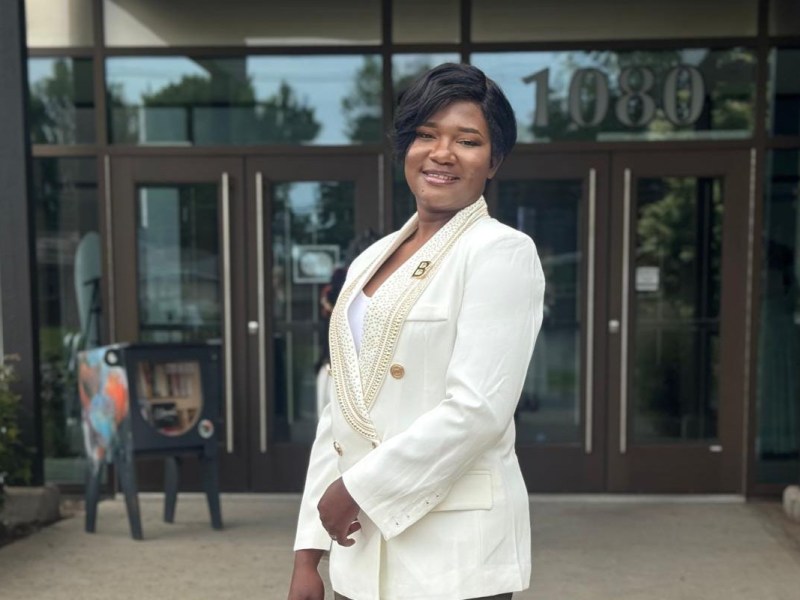A woman stands outside a building entrance, wearing a white blazer, olive green pants, and holding a white bag, smiling at the camera.