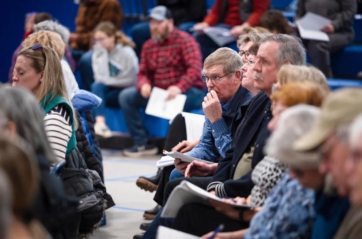 A group of people seated in a gymnasium, attentively listening, with some holding papers.