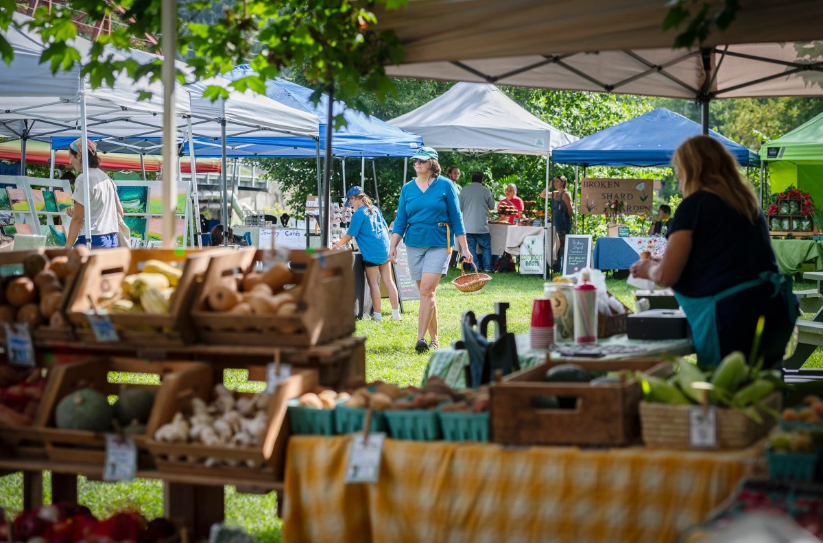 People walking and shopping at an outdoor farmers market with various stalls selling produce and goods under tents.