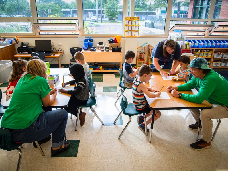 Children and adults sit at tables in a classroom, engaging in various activities and discussions. Shelves with supplies and large windows are visible in the background.