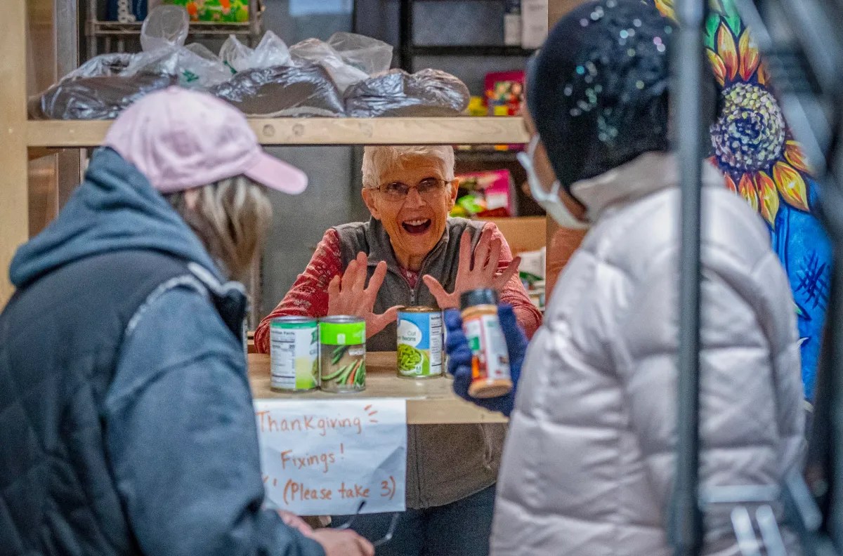 A woman stands behind a counter distributing canned goods to two people. A sign reads, "Thanksgiving fixings! (Please take 3).