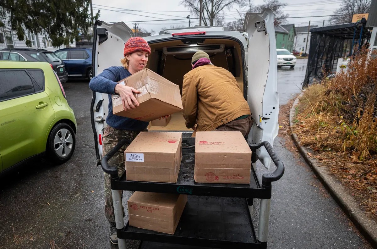 Two people unload boxes from a van onto a cart in a parking lot on a rainy day.