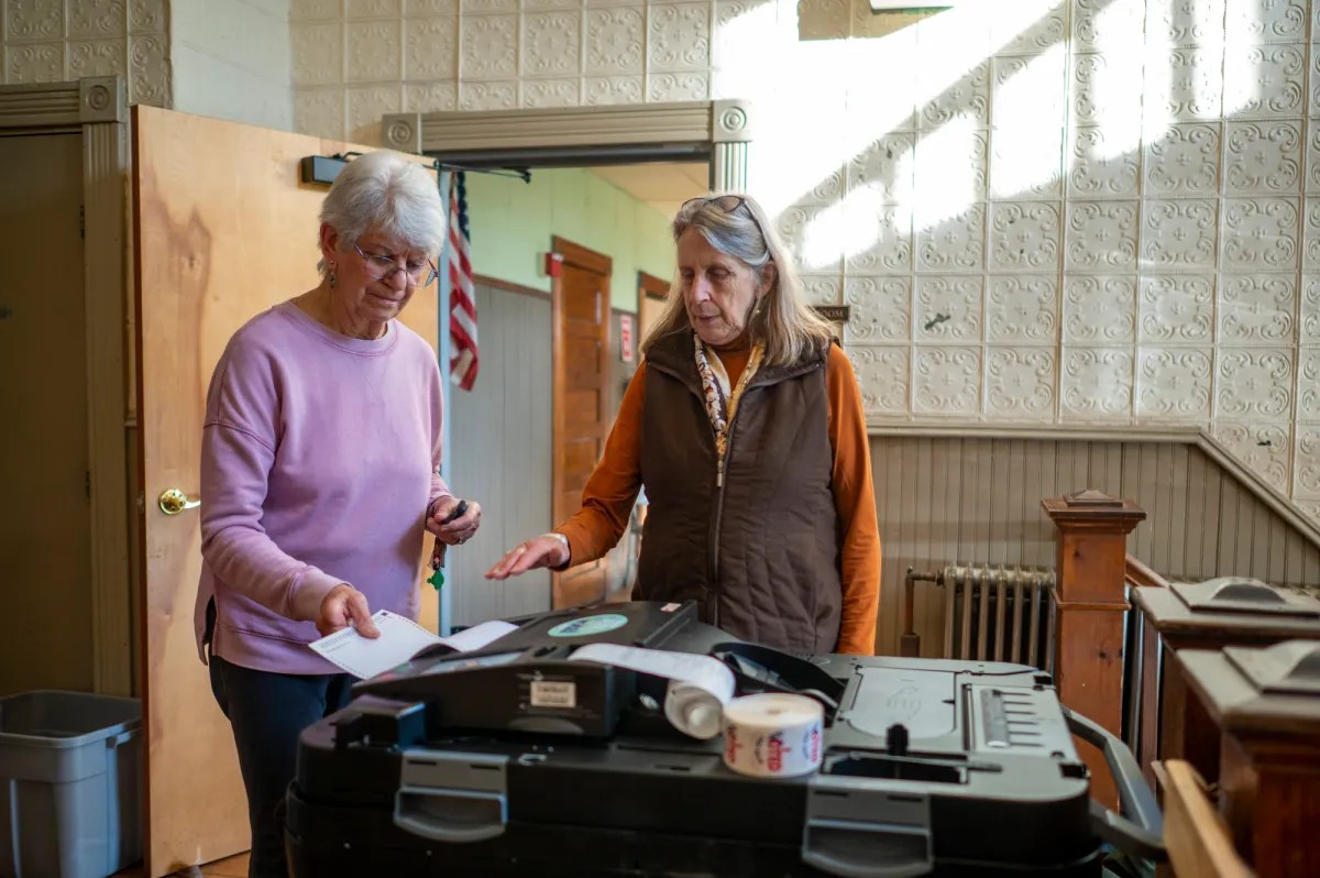 Two women stand next to a voting machine, one inserting a paper into the machine. They are in a room with a wooden door and patterned walls, with sunlight streaming in.