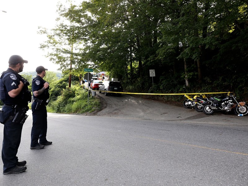Two police officers stand near a road blocked with yellow tape, while several motorcycles and emergency vehicles are parked along the side under trees.