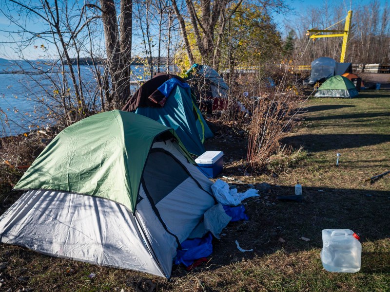 A row of camping tents set up near a river, surrounded by leafless trees. A yellow structure is visible in the background. A plastic water jug and cooler are on the grass.