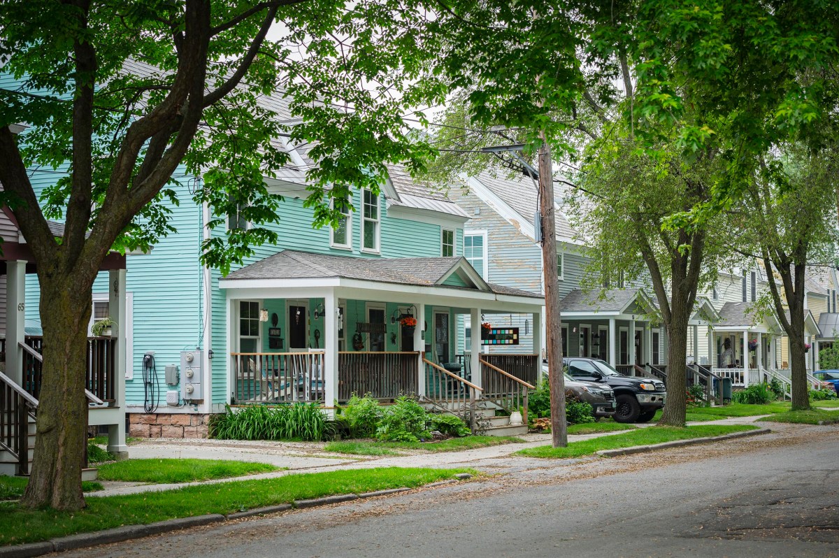 Row of pastel-colored houses with front porches, trees lining the street, and parked cars along the curb in a quiet suburban neighborhood.