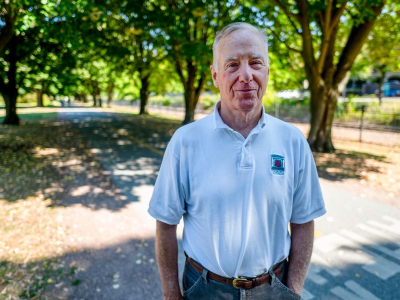 An older man stands on a shaded park path, wearing a white polo shirt and gray pants, with trees and sunlight in the background.