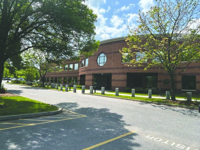 A two-story brick office building with large windows, surrounded by trees and a parking lot under a partly cloudy sky.