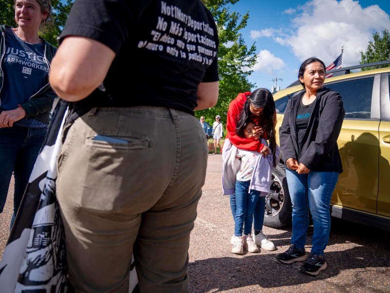 Several people stand next to a yellow car on a sunny day; one person is hugging themselves and looking down, while others stand nearby, some with folded arms.