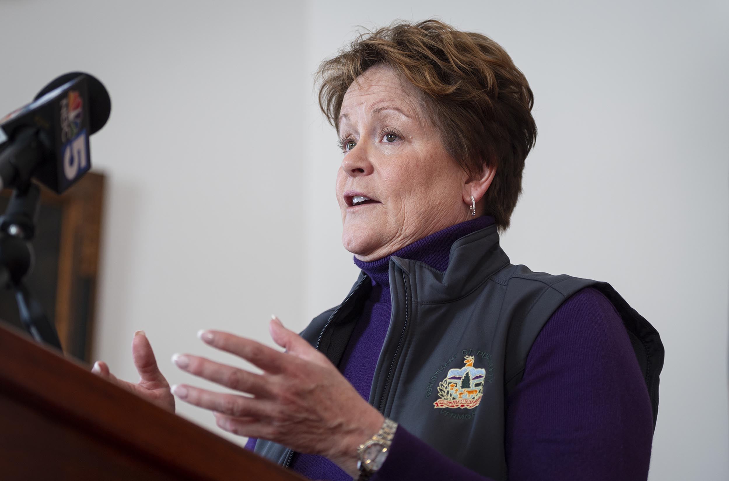 Person in a purple sweater and gray vest speaking at a podium with microphones.
