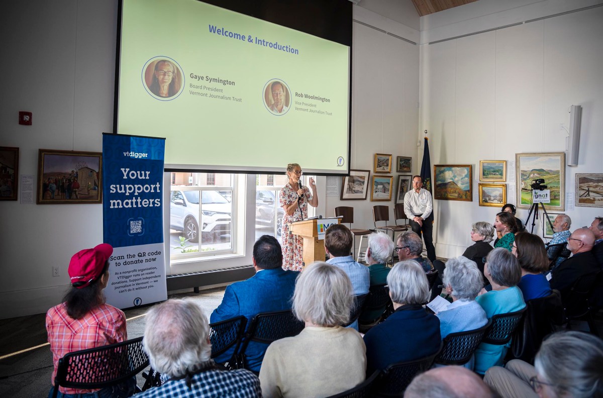 A speaker is addressing an audience in a room decorated with numerous paintings. A large screen overhead shows a "Welcome & Introduction" slide with photos and names of two individuals.