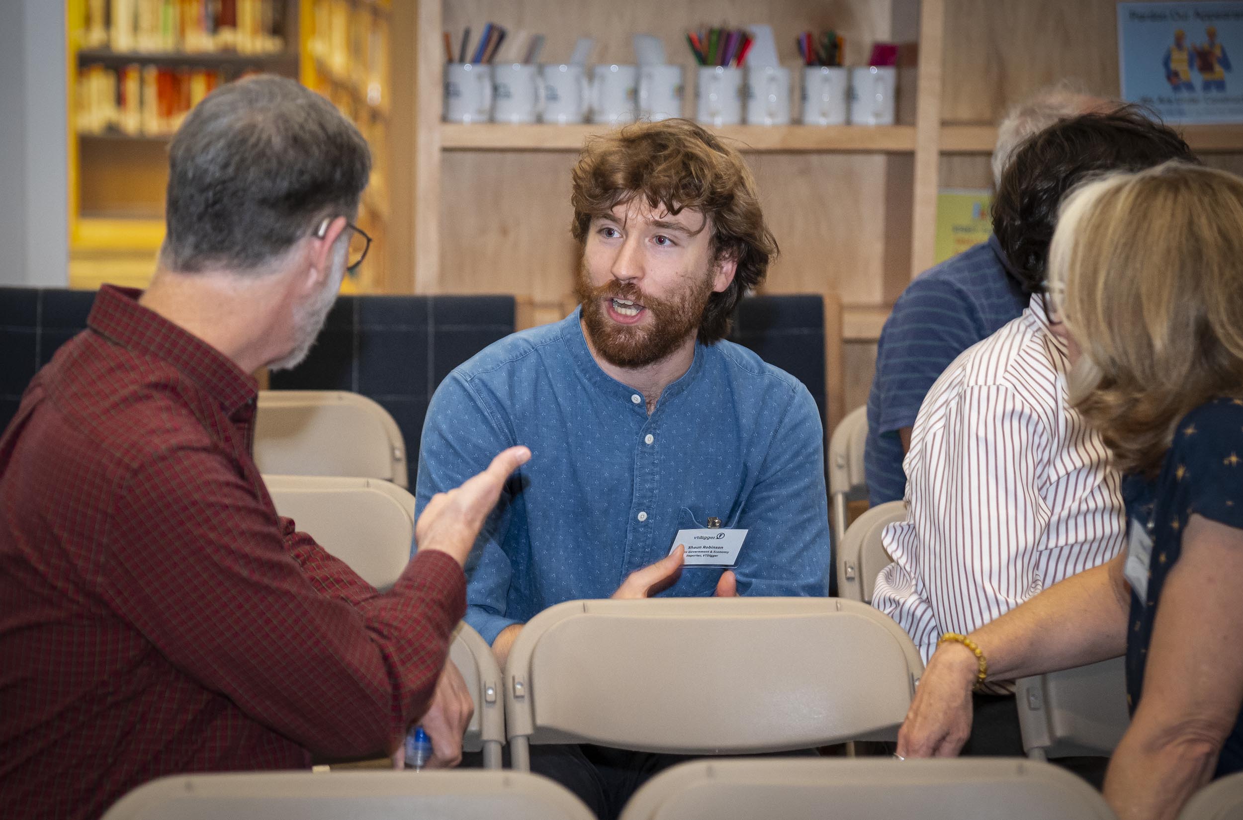 Two men are having a conversation while seated in a room with other people. They appear to be at a meeting or event. Shelves with cups, books, and other items are visible in the background.