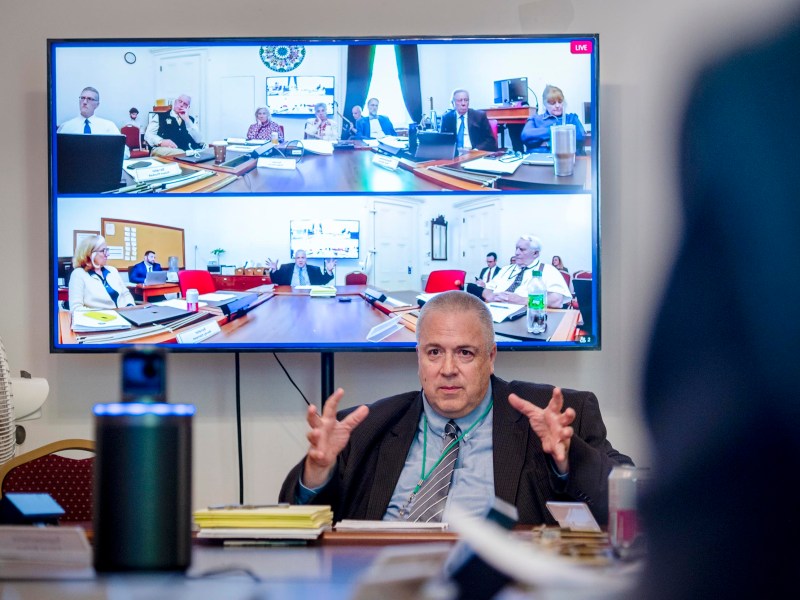 A man sits at a conference table speaking, with a large screen behind him showing multiple people in a video meeting.