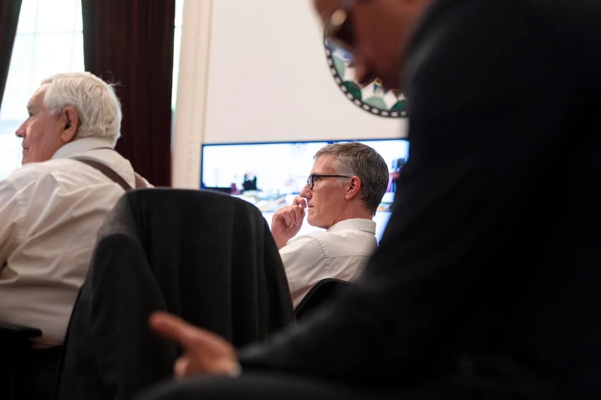 Three men in suits sit in a meeting room, one appears thoughtful, with a monitor and partially visible seal in the background.