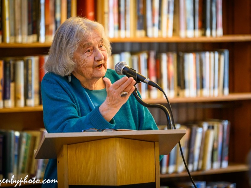 An elderly woman in a teal sweater speaks at a podium with a microphone in front of bookshelves in a library.