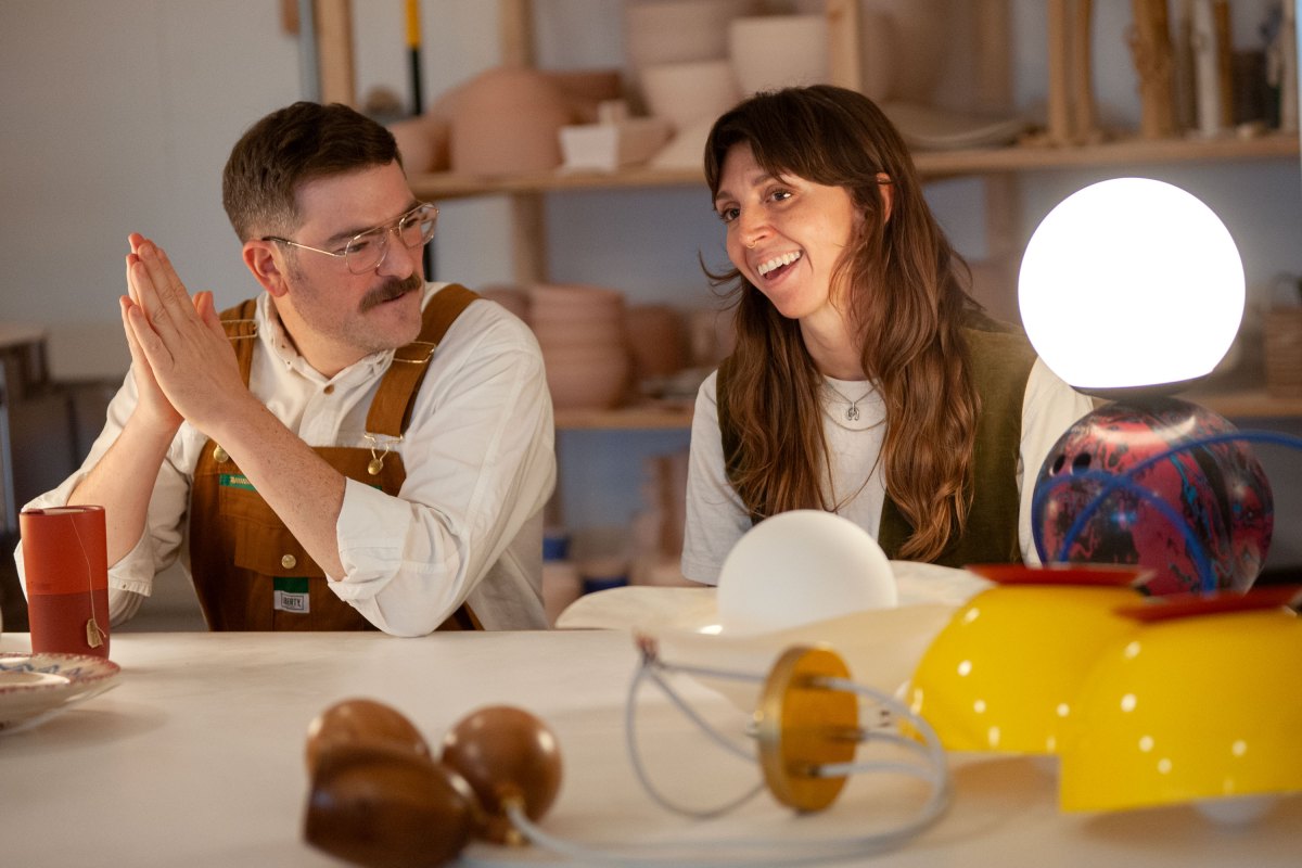 Two people sitting at a table with various colorful objects and a glowing lamp, shelves in the background.