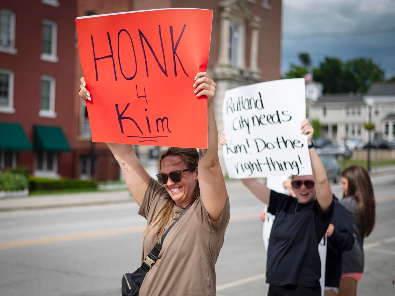 Two people stand on a sidewalk holding protest signs; one reads “Honk 4 Kim” and the other says “Rutland City needs Kim! Do the right thing!”.