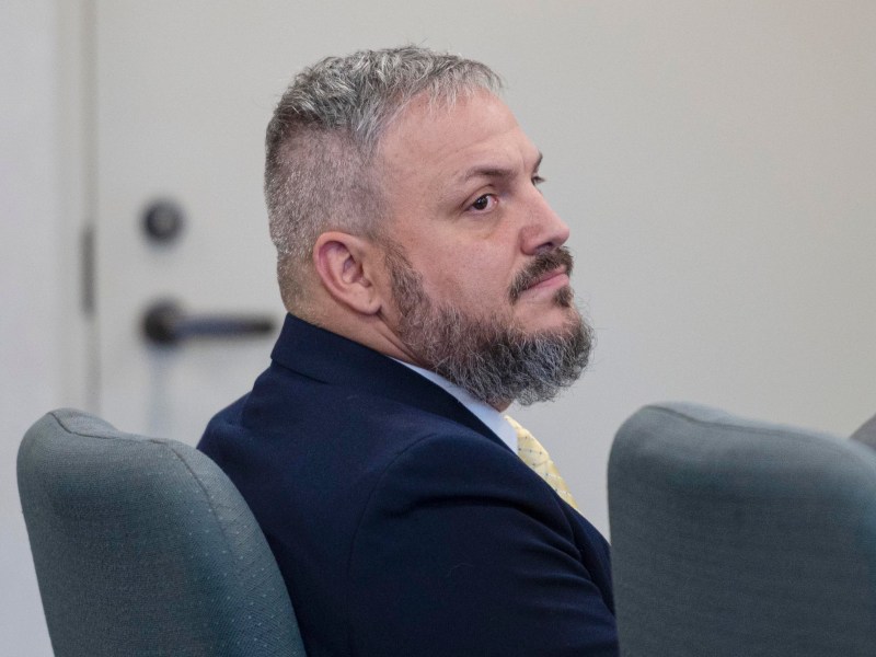 A man with short gray hair and a beard, dressed in a suit and yellow tie, sits in a chair facing slightly to the left in a room with plain walls.