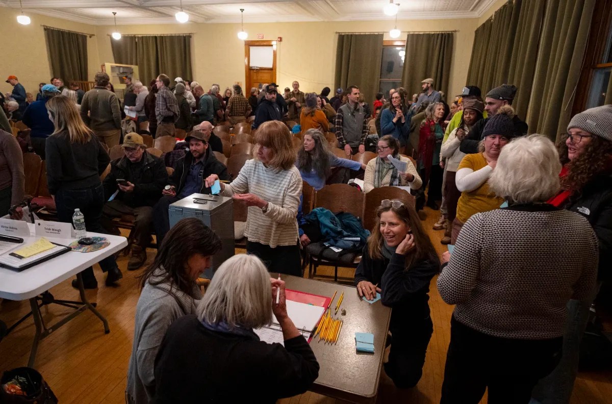 A group of people gather in a room with chairs, engaging in conversations and activities around tables. Some are seated while others stand, with papers and writing materials visible.
