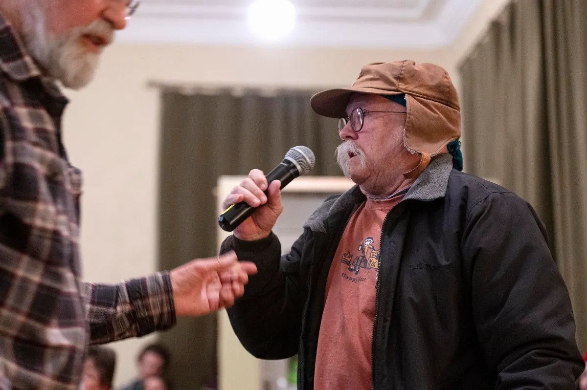 An older person with a mustache in a hat and glasses speaks into a microphone while another person reaches out in front of them.