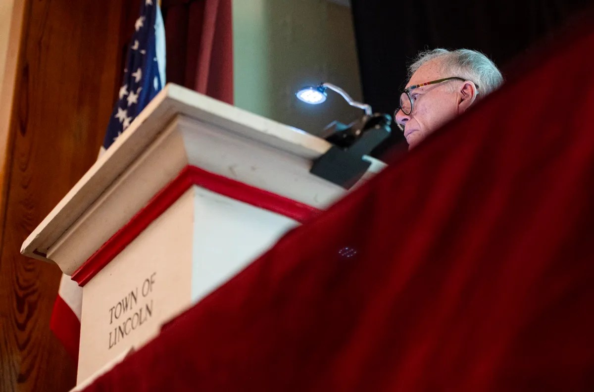 Man standing at a podium labeled "Town of Lincoln" with a reading light, partially obscured by a red curtain.