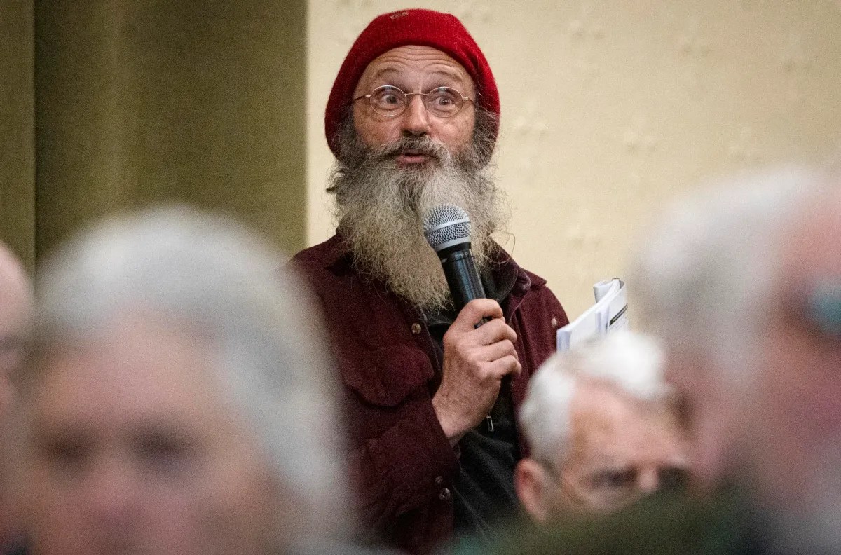 Person with a long beard and red beanie holding a microphone, standing among seated people in a room.