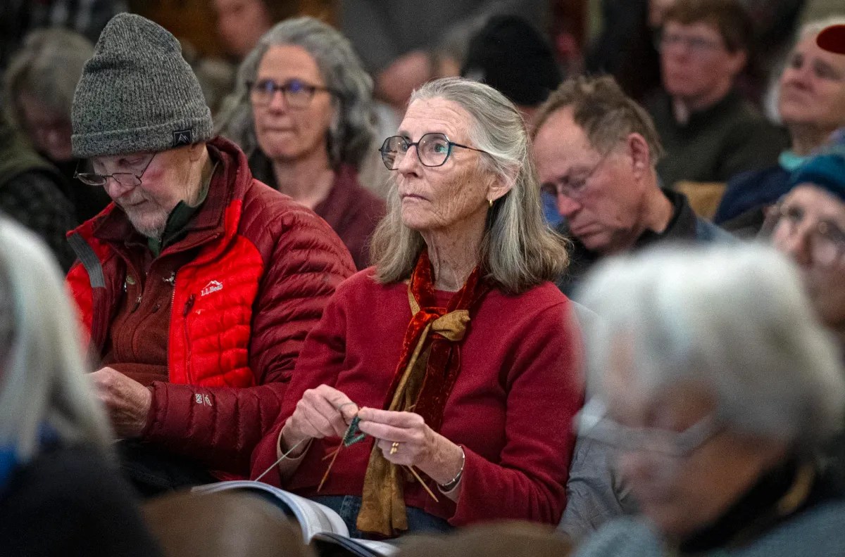 A group of seated older adults attentively listen during a gathering, with one woman knitting.