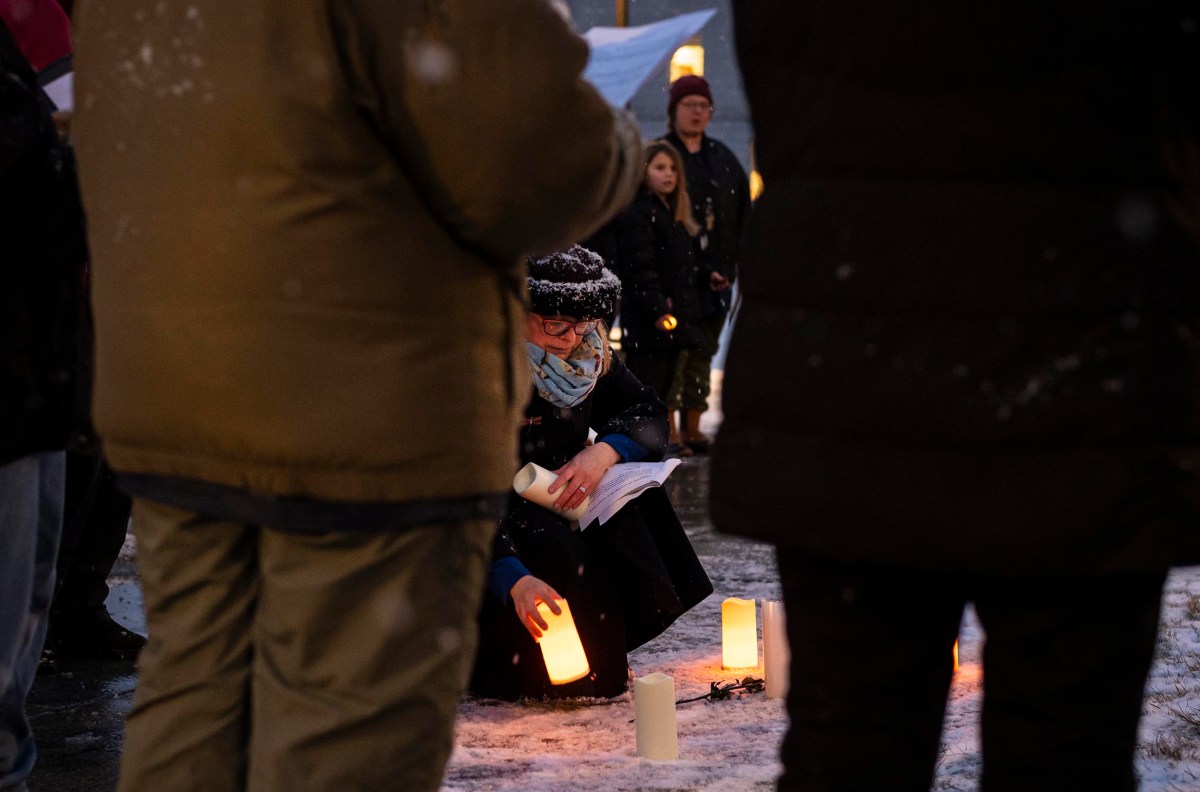 People gather outside in the snow, placing lit candles on the ground, with one person kneeling in the foreground.