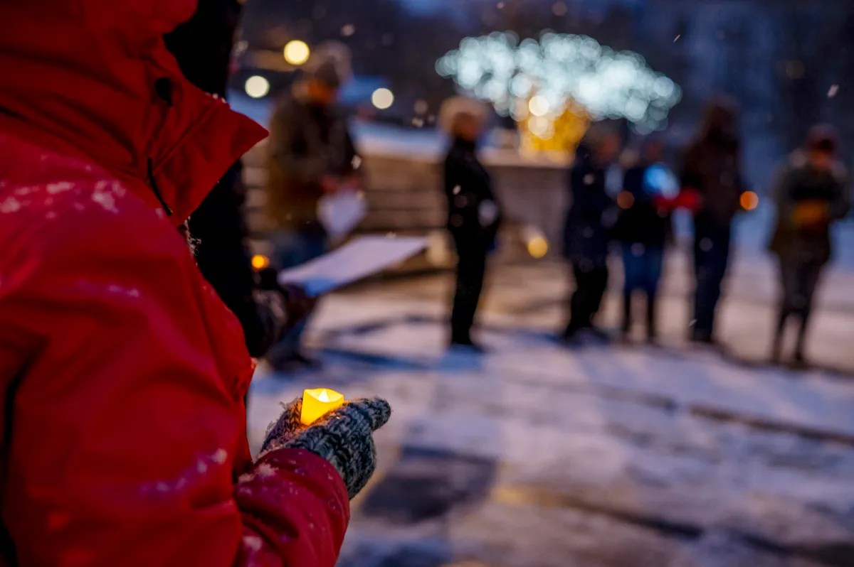 A person in a red coat holds a lit candle in a snow-covered area, with a group of people and trees decorated with lights in the background.