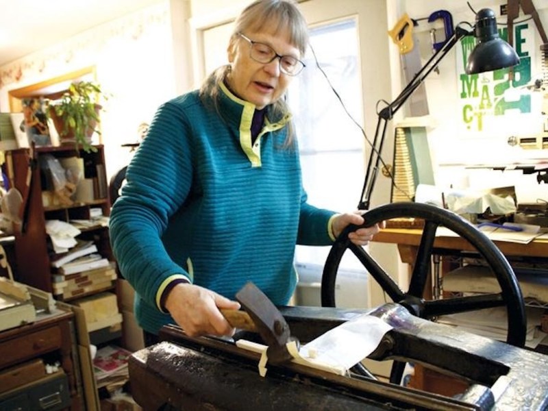 A woman operates a vintage printing press in a cluttered workshop, turning a large wheel and using a tool on a paper or fabric sheet.