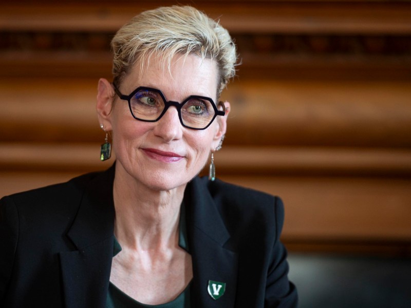A person with short blond hair, glasses, and earrings wearing a dark blazer and a green shirt, seated indoors against a wooden background.
